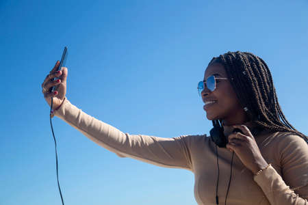 Young black afro woman smiling looking at her cellphone, outdoors with blue sky and sea background. Technology, communication, social networks.の写真素材