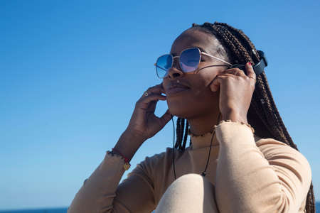 Closeup portrait of young black afro woman smiling with headphones and blue sky background, pretty afro girl transmitting positivity and peace.の写真素材