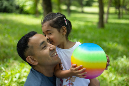 Loving daughter pecking father's cheek, rainbow ball nestled between them amid verdant park landscapeの写真素材