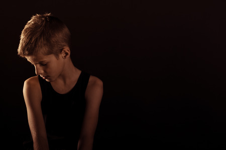 Lonely Slim Boy in Sleeveless Shirt Looking Down Against Dark Background with Copy Space on the Left Side.の写真素材