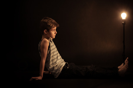 Thoughtful Boy Sitting on the Floor and Leaning his Back Against his Arms, Looking Straight at Glowing Light Bulb on Stand.の写真素材