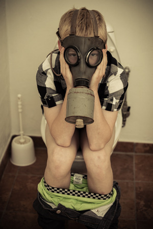 Single male child in gas mask sitting on toilet in bathroom with pants down and hands on faceの写真素材