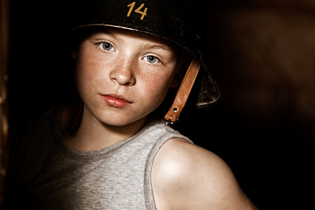 Serious little boy playing soldier in the shadows while wearing large army helmet with number 14 painted on itの写真素材