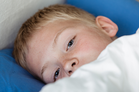 Close up of cute little blond boy under white blanket in blue bed waking up with open eyesの写真素材