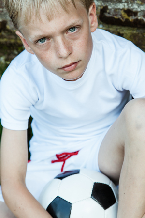 Close up of blond and blue eyed boy wearing all white crouching with soccer ballの写真素材