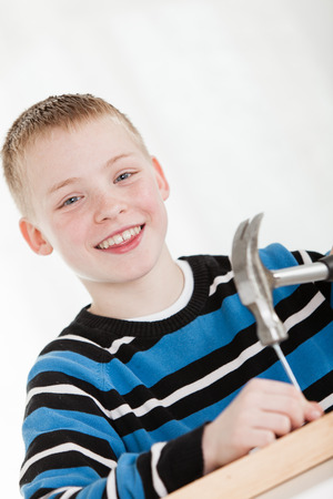 Smiling boy wearing blue and black sweater hammers nail into piece of wood against a white backgroundの写真素材