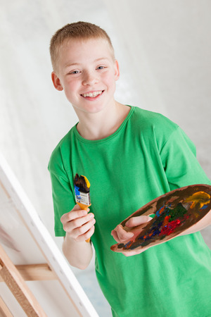 Boy in green shirt holding painters pallet and brush near easel with canvas against a light backgroundの写真素材