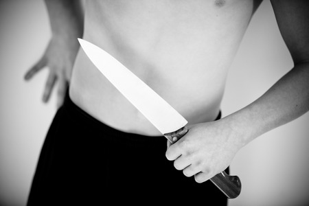 Cropped and angled close up view on boy holding large butcher knife near torso with hand leaning against wallの写真素材