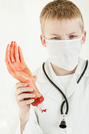 Single blond boy dressed as surgeon with white face mask holding mock dismembered and bloodied handの写真素材