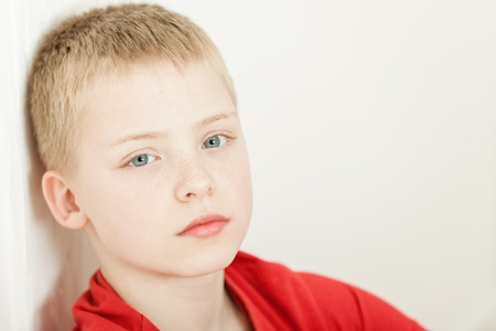 Single sick little blond boy wearing red shirt with tired expression sitting back against wall with copy spaceの写真素材