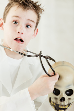 Shocked young child dressed up as a doctor in white lab coat checking human skull with stethoscopeの写真素材
