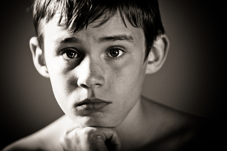 Black and White Head and Shoulders Close Up of Young Shirtless Teenage Boy Staring at Camera with Worried Expression and Chin Resting on Hand in Vignette Studio with Dark Background and Copy Spaceの写真素材
