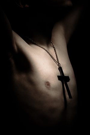 Lighted bare chest of boy wearing large chain and crucifix symbol with arms up in dimly lit areaの写真素材