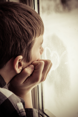 Partial side view of unidentifiable teenage boy looking out window with hand on side of face toward heart symbol shape written on glassの写真素材