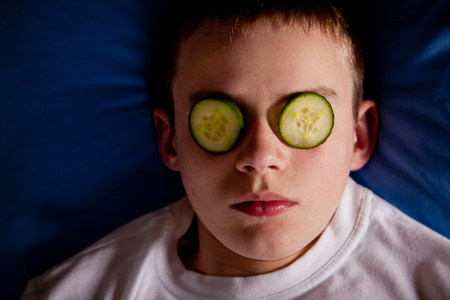 Boy in bed with cucumber slices over his eyes while he lays his head on a blue pillow and wearing a white shirtの写真素材