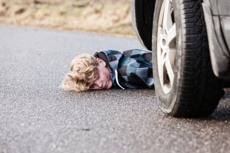 Close Up of Car Accident Fatality - Young Teenage Boy Has Been Hit by a Car and is Lying Unconscious on Road Pavement with Various Injuries, Car Visible in Foregroundの写真素材