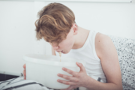 Sick or drunk young boy vomiting into a white bowl as he sits on his bed at home, close up realistic viewの写真素材