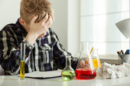 Teenage Boy Working on Chemistry Homework with Head in Hands and Sitting at Desk Surrounded by Beakers with Colorful Liquidsの写真素材