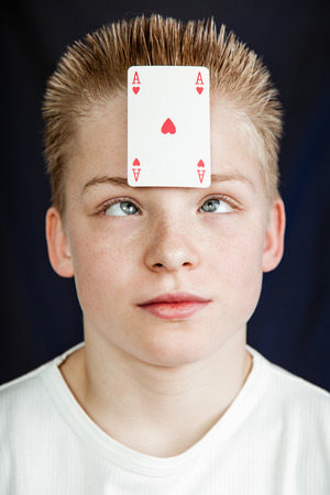 Head and Shoulders Close Up of Pre Teenage Boy with Spiked Blond Hair Looking Cross Eyed at Ace of Hearts Playing Card Stuck to Forehead in Studio with Dark Backgroundの写真素材