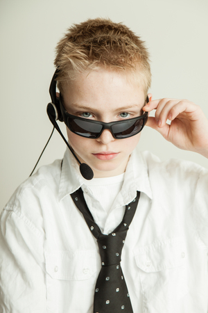 Occupation Concept Image - Head and Shoulders Portrait of Young Blond Pre Teenage Boy with Spiked Hair Dressed as Businessman and Wearing Head Set, Tilting Sunglasses Downの写真素材