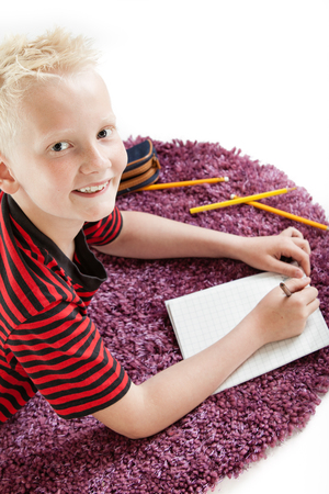 Attractive friendly young boy lying on a rug with a blank sheet of paper and pencils looking up at the camera with a warm smile, isolated on whiteの写真素材