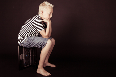 Pensive boy rest hands on knees while sitting on keyboard box and wearing stripped shirtの写真素材