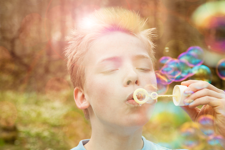 Young boy blowing soap bubbles outdoors in a garden or woodland with a bright sun flare back lighting him and iridescent bubbles floating in the airの写真素材