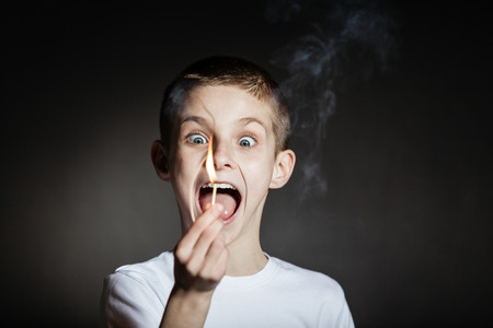 Frightened boy wearing white shirt with eyes and mouth wide open holding lit match in darkened roomの写真素材