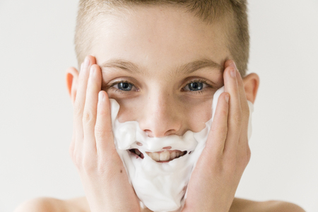 Close Up Head and Shoulders Portrait of Smiling Young Boy Applying Shaving Cream to Face in Studio with White Background - Boy Playing Grown Up and Looking Happyの写真素材