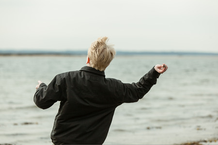 Rear view of single blond youth in dark jacket throwing something toward lake. Background out of focus.の写真素材