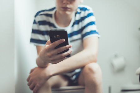 Crop view of incognito boy teenager sitting on toilet seat pantless with at smartphone in his handの写真素材