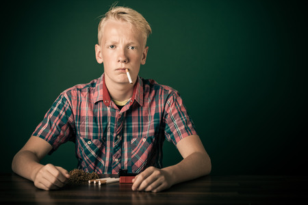 Young teenage boy sitting smoking at a dining table with the cigarette dangling from his mouth and a defiant expressionの写真素材