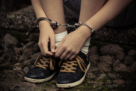 Young boy in handcuffs and sneakers sitting with his hands resting on his shoes on old cobblestones after being arrested, close up view of the manaclesの写真素材