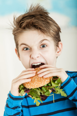 Young boy biting into a salad burger with fresh rocket leaves on a sesame bun with a look of surprise and determinationの写真素材