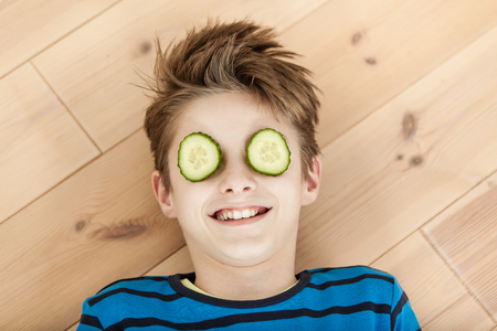 Laughing young boy with two slices of fresh cucumber on his eyes lying on a wooden floor conceptual of a beauty regimeの写真素材