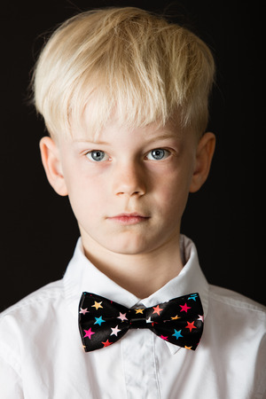 Handsome little blond boy in a formal white shirt and smart bow tie decorated with colorful stars on black looking directly at the camera in a close up head shotの写真素材