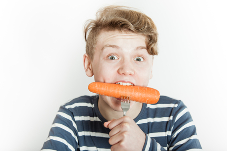 Excited male teenager in striped blue and white shirt biting into large carrot with fork stuck in the middle.の写真素材