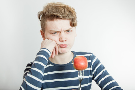 Annoyed male child in long sleeve blue and white shirt with hand on cheek and fork stuck in red appleの写真素材