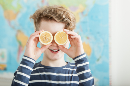 Cute smiling boy with wavy hair holding lemon halves that cover both eyes while standing in front of world mapの写真素材