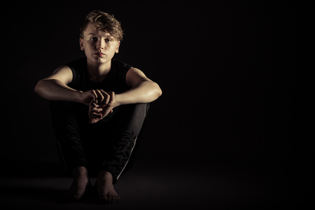 Sad barefooted child seated with arms around knees and copy space beside him over dark backgroundの写真素材