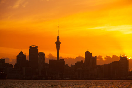 Auckland, New Zealand skyline at sunset with vibrant orange sky and silhouetted buildings.