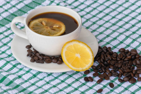 Coffee in white cup with roasted coffee beans, and lemon, on beautiful pattern tablecloth, is background.の写真素材