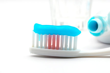 Blue toothpaste on toothbrush bristles, toothpaste tube, and a glass of water on white background.