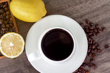 Top view of warm black coffee in white cup with yellow lemon, coffee beans, and lemon slices on vintage brown wooden background. Healthy drink concept.の写真素材