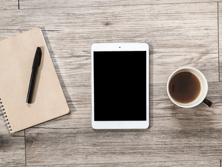 White Tablet with blank black screen and a cup of coffee and notebook with pen on the wooden backgroundの写真素材