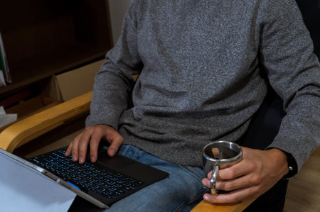 Young man holding laptop in lap, working on it, typing in a grey pullover with a coffeeの写真素材