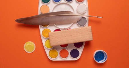 Watercolor paints and a feather on an orange background, top viewの写真素材