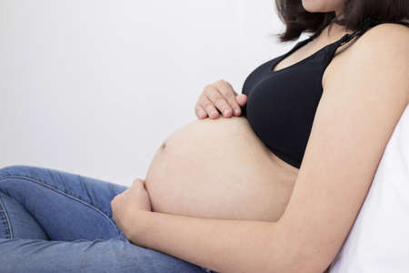 Happy pregnant asian woman sitting on bed and touching her big bellyの写真素材