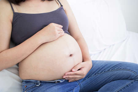 Pregnant asian woman sitting on bed and touching her big belly