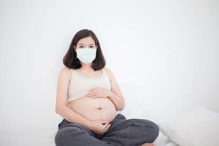 Pregnant asian woman in medical face mask to protect Covid-19 (Coronavirus) and PM 2.5 sitting on...